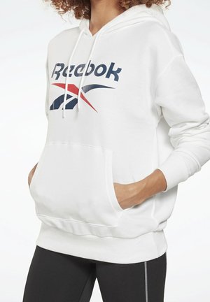 Person iført en hvid Reebok hættetrøje med forlomme og snore, kombineret med sorte leggings med hvide syninger.