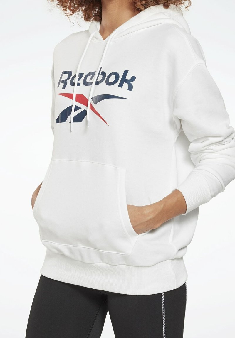 Personne portant un sweat à capuche blanc Reebok avec poche avant et cordons, associé à des leggings noirs avec coutures blanches.
