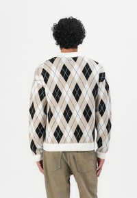 Beige argyle sweater with black and white diamonds, ribbed white cuffs and hem, worn over light brown pants. Logo tag at the back neckline.