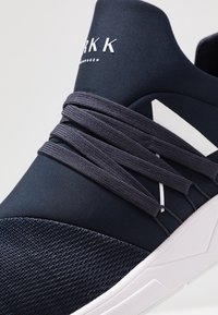 Marinblå atletikskor med en texturerad stickad ovandel, elegant design, elastiska snören och en vit logotyp. Sulan är vit och strukturerad.