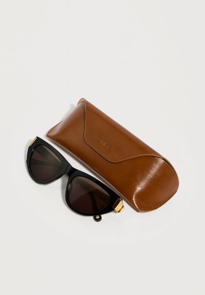 Μαύρα γυαλιά ηλίου σε σχήμα γάτας με χρυσές λεπτομέρειες δίπλα σε καφέ δερμάτινη θήκη προστασίας σε λευκό φόντο.