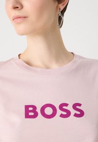 Šviesiai rožinė medvilnės marškinėliai su ryškiai purpuriniu "BOSS" užrašu. Apvali iškirptė ir glotni medžiagos tekstūra, su artimu dizaino vaizdu.