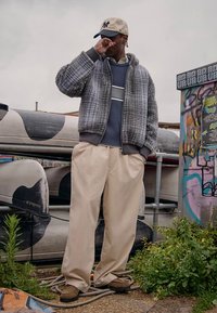 Grijze bomberjack met ruitpatroon, blauw crewneck met strepen, beige wijde broekspijpen en bruine schoenen. Casual streetwearstijl.