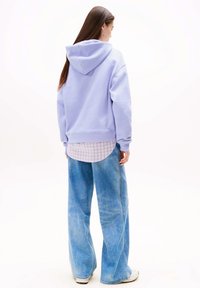 Lavendelkleurige capuchontrui met een losse pasvorm, gedragen over een geruit overhemd. Gecombineerd met wijde blauwe spijkerbroeken. Casual stijl.