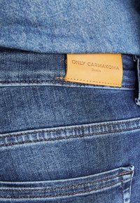 Vaqueros de mezclilla con un primer plano del bolsillo trasero y una etiqueta de cuero color marrón claro que dice "ONLY CARMakoma Denim". Tejido azul con costuras en contraste.