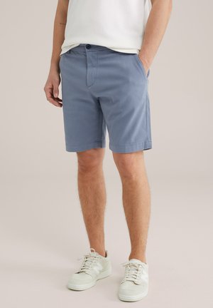 Pantaloni corti in cotone azzurro chiaro con una finitura testurizzata, dotati di un bottone frontale e tasche laterali, abbinati a scarpe da ginnastica bianche.