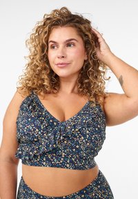 Kiharatukkainen nainen sinisessä kukkakuviollisessa röyhelöisessä lyhyessä paidassa, poseeraa koskettaen hiuksiaan, neutraali tausta.