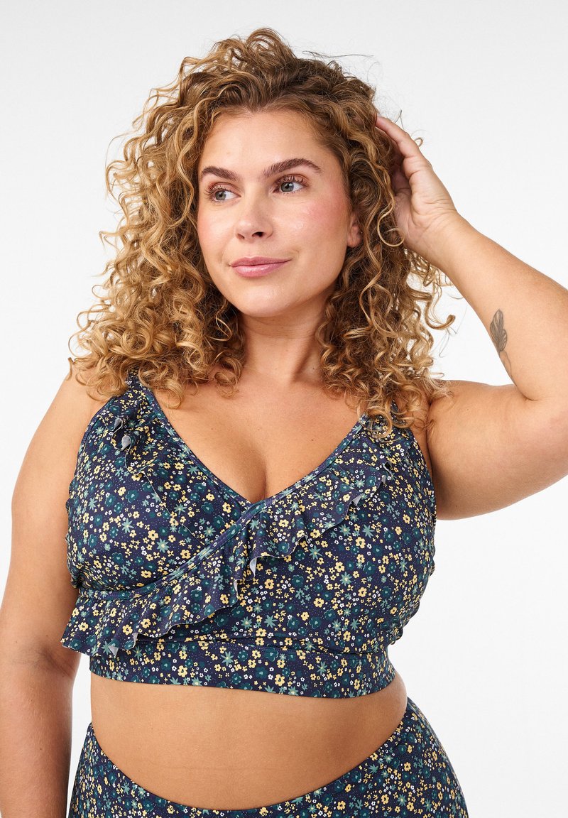 Kiharatukkainen nainen sinisessä kukkakuviollisessa röyhelöisessä lyhyessä paidassa, poseeraa koskettaen hiuksiaan, neutraali tausta.