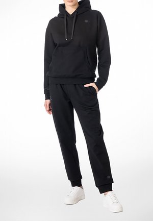 Henkilö pukeutuneena mustaan hupulliseen svetariin ja sopiviin mustiin collegehousuihin, seisoo käsi yhdessä taskussa ja valkoiset lenkkarit jalassa.