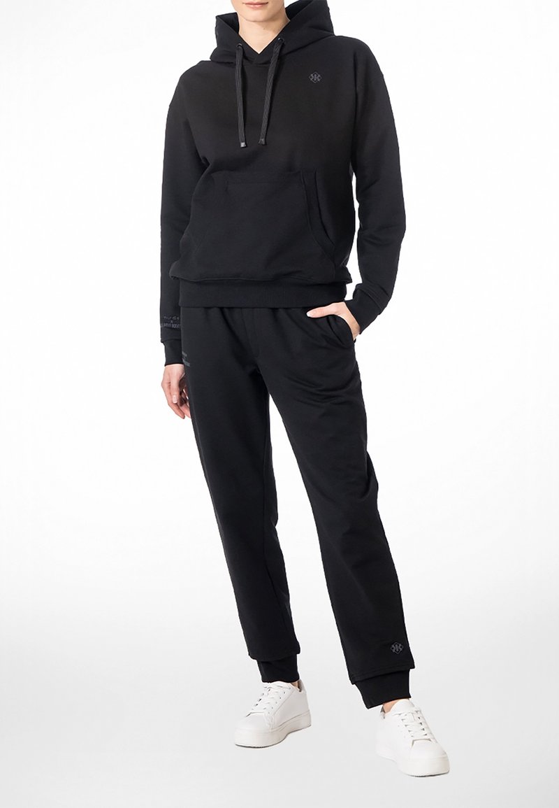 Person trägt schwarzen Kapuzenpullover und passende schwarze Jogginghose, steht mit einer Hand in der Tasche und weißen Turnschuhen.