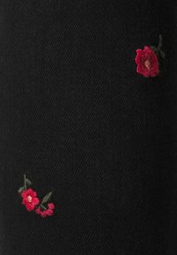 Tela negra con dos pequeñas flores rojas bordadas y hojas verdes, mostrando textura y detalles de costura finos.