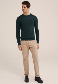 Donkergroene gebreide trui met ronde hals, beige chinos en zwarte veterschoenen. De stof lijkt zacht met een gestructureerde afwerking.