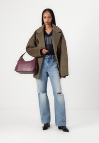 Mujer con abrigo marrón oversize, camiseta de rayas azul marino, vaqueros en azul claro desgarrados, botas negras y un bolso grande burdeos.