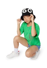Camiseta verde con un bolsillo brillante, combinada con shorts de mezclilla. Sombrero tipo cubo negro con letras blancas, zapatillas blancas y calcetines de tobillo.