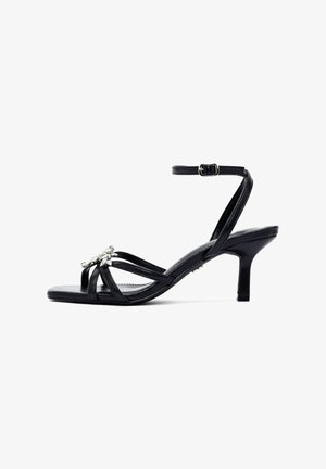 Black heeled sandals with a strappy design, featuring a small buckle and jeweled embellishment on the front. Smooth leather texture.