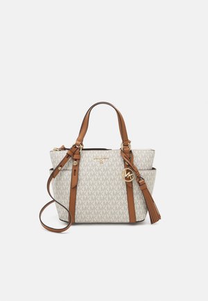 Sac cabas beige avec anses en cuir marron, motif monogramme Michael Kors, breloque logo dorée et détails à pompons sur fond blanc.