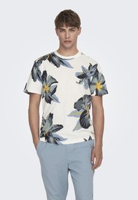 Kortermet kremfarget t-skjorte med blomstermønster i blått, gult og svart. Sammen med lyseblå bukser står modellen med hendene i lommmene.