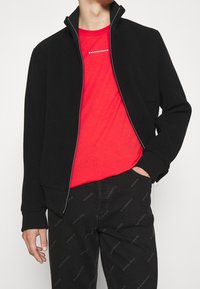 Giacca nera con zip, maniche e colletto a costine, sopra una t-shirt rossa. Pantaloni neri con lettering "PARIS" ripetuto in un motivo sottile.