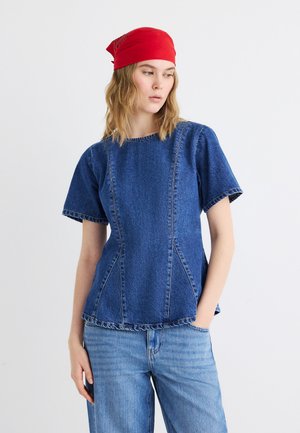 Femme portant un foulard rouge, un haut bleu en denim à manches courtes et un jean bleu clair, debout avec une main dans la poche devant un fond uni.