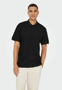 Sort polo shirt med krave og tre knapper, korte ærmer, ensartet tekstur, båret med lyse bukser. Simpelt og afslappet design.