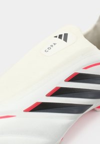 Λευκό ποδοσφαιρικό παπούτσι με λεία υφή, μαύρες και κόκκινες λεπτομέρειες, και έναν προεξέχοντα λογότυπο στο πλάι με σύγχρονο σχεδιασμό.