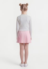 Dívka se světlými vlasy upravenými do dvojitých drdolů a copánků, oblečená v bílém topu s dlouhým rukávem, růžové plisované sukni a bílých teniskách, stojící zády.