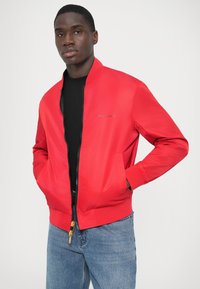 Rød bomberjakke med lynlås foran, to sidelommer, ribbet manchetter og en logodetalje på brystet, kombineret med blå jeans.