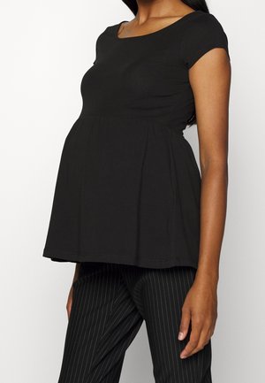Femme portant un haut de maternité noir à manches courtes, col rond et coupe ample, sur un pantalon noir à fines rayures.