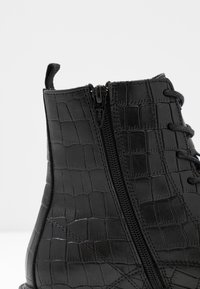 Bottine noire à cheville avec une tige en cuir à motif crocodile, un laçage à l'avant et une fermeture éclair externe pour un accès facile.