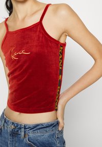 Top corto in velluto rosso con spalline sottili, caratterizzato da un logo dorato e una pannellatura laterale nera accentuata da scritte gialle.