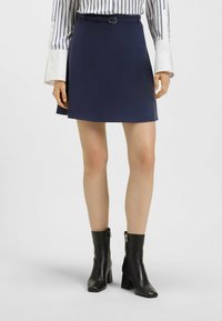 Granatowa spódnica w kształcie litery A z paskiem z srebrną klamrą, w zestawie z czarnymi botkami za kostkę. Materiał jest gładki, z minimalną teksturą.