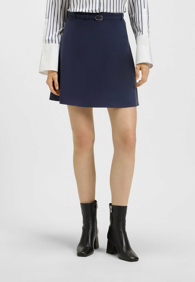 Granatowa spódnica w kształcie litery A z paskiem z srebrną klamrą, w zestawie z czarnymi botkami za kostkę. Materiał jest gładki, z minimalną teksturą.