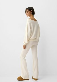 Cream, oversized knit sweatshirt with a wide neckline, paired with matching fitted flared pants and tan slip-on shoes. Minimalist design.