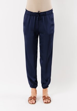 Pantaloni jogger in satin blu navy con elastico in vita e cordoncino regolabile, con orli a punta e una texture liscia. Abbinati a sandali marroni.