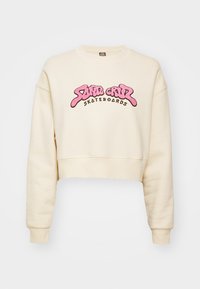 Felpa accorciata color crema con collo e polsini a coste. Grande logo rosa "Santa Cruz Skateboards" sulla parte anteriore. Texture morbida.