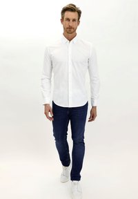 Camicia bianca con colletto, indossata con jeans blu navy e sneaker bianche. Il tessuto appare liscio; la vestibilità è sagomata.