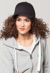 Zwarte baseballpet van stof met een gebogen rand, gedragen met een grijze hoodie met een witte rits en groene logo-accenten.