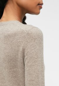 Pull en maille beige clair avec une texture douce, encolure ronde et manches montées, présentant une coupe près du corps et un design minimaliste.