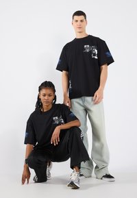 Black graphic t-shirts feature bold white and blue designs. The male model wears light denim, while the female model pairs with black pants.
