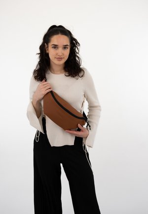 Brun syntetisk crossbody taske med en sort lynlås, slank form og justerbar rem, holdt af en person iført en lys sweater.
