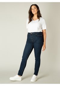 Donkerblauwe high-waisted jeans met een smalle pasvorm, voorzakken, sluiting met één knoop en contrasterende stiksels, gecombineerd met een wit T-shirt en witte sneakers.