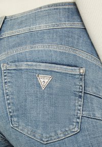 Lyseblå denim jeans med et trekantet Guess-logo patch på baglommen, med subtile syninger og teksturdetaljer.