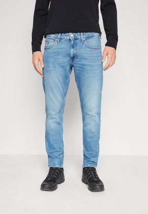 Jeans de mezclilla azul claro con corte ajustado, que presentan un ligero desgaste, diseño de cinco bolsillos y sutiles detalles de costura. Llevados con zapatos negros.