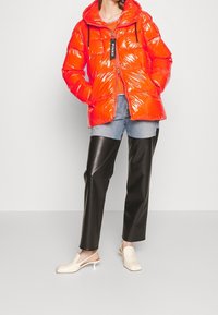 Jasnopomarańczowa kurtka puchowa o błyszczącym wykończeniu, z kapturem, zapinana na zamek, z sznurkami, zestawiona z czarnymi skórzanymi spodniami i beżowymi butami.