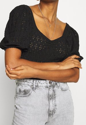 Persona que lleva una camiseta negra con mangas abullonadas y ojales y unos vaqueros de tiro alto gris claro con los brazos cruzados sobre el pecho.