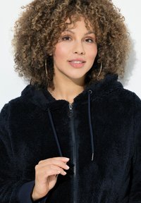Kvinde med krøllet hår og hoops øreringe justerer snoretræk på en navy blå fuzzy zip-up hættejakke mod hvid baggrund.