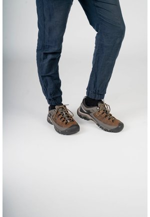 Ben iført mørkeblå joggingbukser og brune vandrestøvler med sorte såler, stående på en ren hvid overflade.