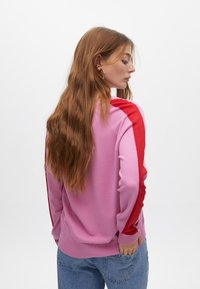 Pullover aus weichem Stoff, mit pinkem Oberkörper und roten Schulterakzenten. Das Design umfasst gerippte Bündchen und Saum sowie eine lockere Passform.