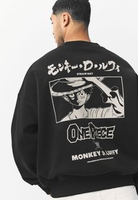 Sort sweatshirt med stort billede af en karakter med en stråhat, ledsaget af japansk tekst og "ONE PIECE"-logoet nedenfor.