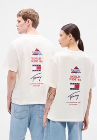 T-shirts brancas de algodão com uma estampa gráfica nas costas. O texto inclui "WORLD-WIDE '85" e "ROUPA PARA O EXTERIOR", com várias cores usadas.